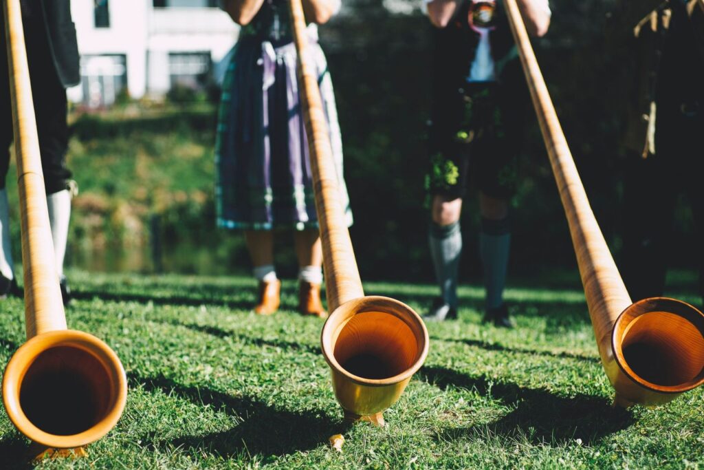 Three musicians in traditional Swiss attire playing wooden alphorns on a grassy field.